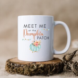 Puede incluir: Taza de cerámica blanca con una calabaza turquesa y un diseño floral. El texto en la taza dice "Meet me at the Pumpkin Patch".