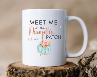 Può includere: Una tazza in ceramica bianca con una zucca turchese e un motivo floreale. Il testo sulla tazza recita "Meet me at the Pumpkin Patch".