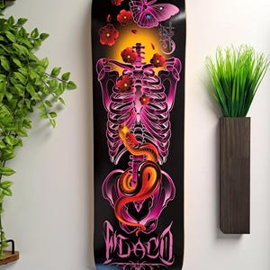 May include: A black skateboard deck with a pink and red skeleton design. The skeleton is surrounded by flowers and a butterfly. The words "FLACO" are printed on the bottom of the deck.