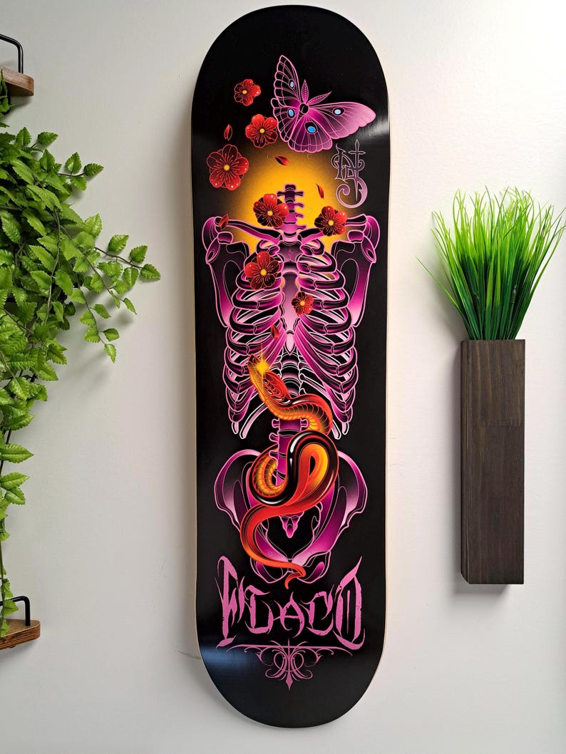 May include: A black skateboard deck with a pink and red skeleton design. The skeleton is surrounded by flowers and a butterfly. The words "FLACO" are printed on the bottom of the deck.