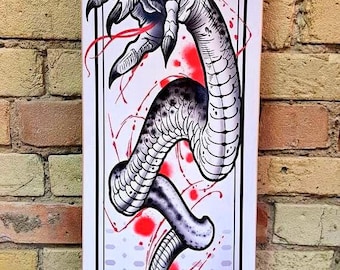May include: A white skateboard deck featuring a black and white illustration of a dragon's claw and tail. The dragon is surrounded by red splatters of paint. The deck has a black and white pattern at the bottom and a red symbol in the centre.