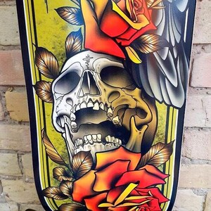 May include: A black and yellow skateboard deck with a traditional tattoo style design. The design features a skull with an open mouth, two red roses, and a grey bird wing.