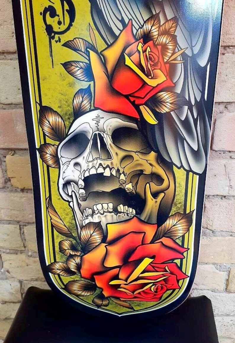 May include: A black and yellow skateboard deck with a traditional tattoo style design. The design features a skull with an open mouth, two red roses, and a grey bird wing.