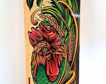 Könnte beinhalten: Ein Skateboard-Deck mit einer detaillierten Illustration eines Hahns mit grünen Federn, rotem Kamm und roten Kehllappen. Das Design umfasst goldene Weizenähren und florale Ornamente auf hellem Holzuntergrund mit dem Schriftzug "LOCO" in Rot.