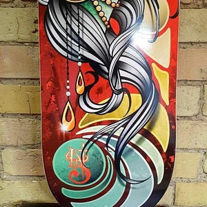 Pu&ograve; includere: Un deck da skateboard longboard con un design colorato che presenta il viso di una donna, i suoi capelli e un cerchio decorativo con un motivo a goccia. Il deck ha uno schema di colori verde, giallo e rosso con un motivo a spirale nella parte inferiore.