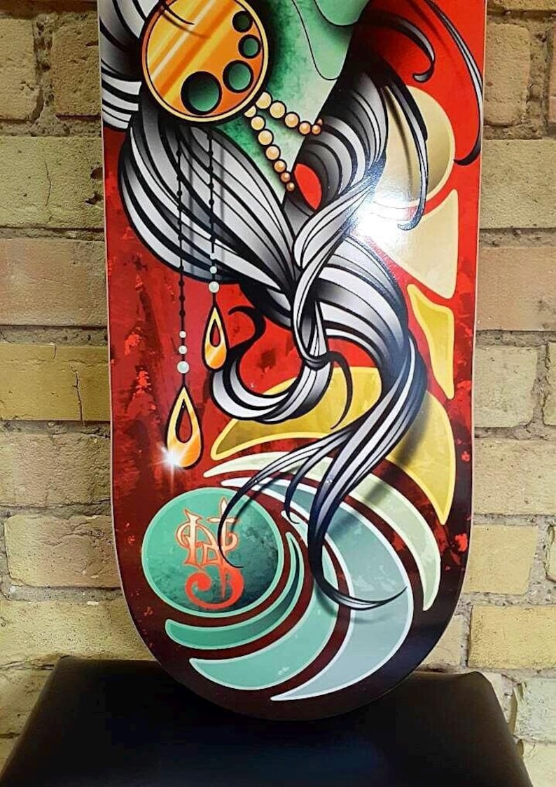 Pu&ograve; includere: Un deck da skateboard longboard con un design colorato che presenta il viso di una donna, i suoi capelli e un cerchio decorativo con un motivo a goccia. Il deck ha uno schema di colori verde, giallo e rosso con un motivo a spirale nella parte inferiore.