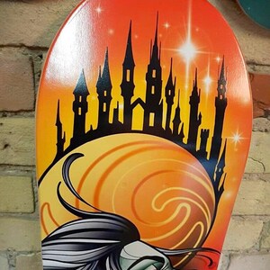 Pu&ograve; includere: Un mazzo di skateboard colorato con una silhouette nera di un castello su uno sfondo di tramonto. Un viso di donna con un design a spirale arancione e giallo &egrave; in primo piano.