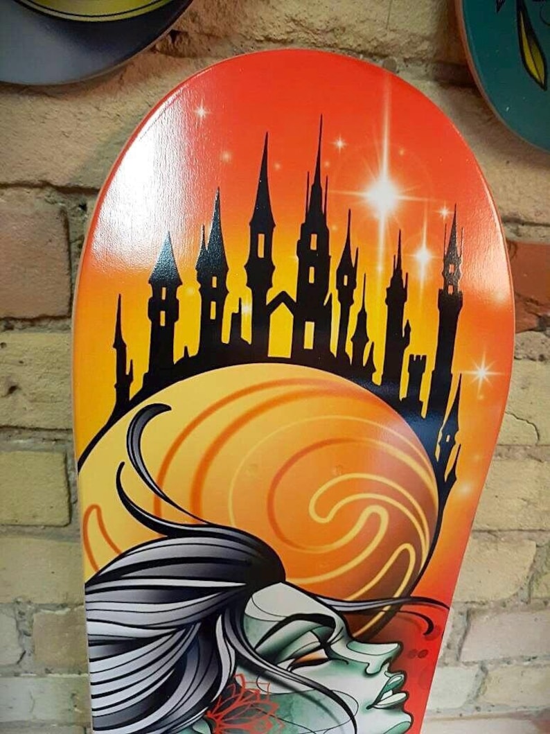 Pu&ograve; includere: Un mazzo di skateboard colorato con una silhouette nera di un castello su uno sfondo di tramonto. Un viso di donna con un design a spirale arancione e giallo &egrave; in primo piano.