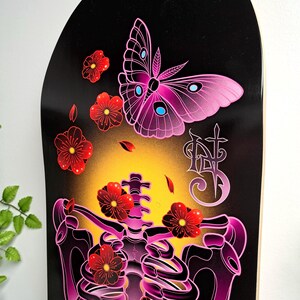 May include: A black skateboard deck with a pink and purple skeleton with red flowers and a pink butterfly. The butterfly has blue accents. The deck has a yellow glow around the skeleton.