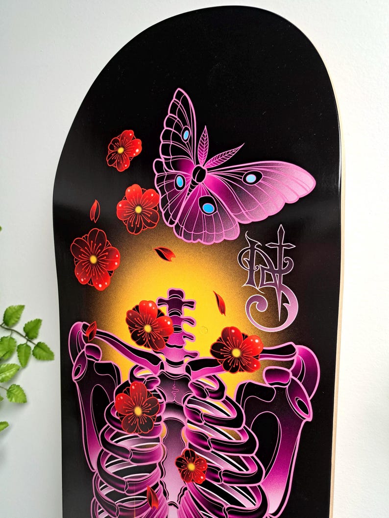 May include: A black skateboard deck with a pink and purple skeleton with red flowers and a pink butterfly. The butterfly has blue accents. The deck has a yellow glow around the skeleton.