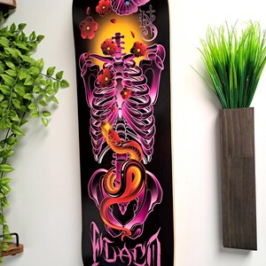 May include: A black skateboard deck with a pink and purple skeleton design. The skeleton is surrounded by pink flowers and a butterfly. The word "FLACO" is written in pink at the bottom of the deck.