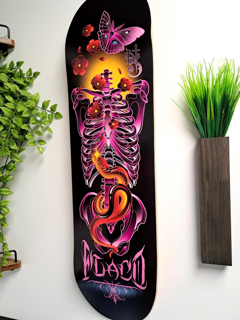 May include: A black skateboard deck with a pink and purple skeleton design. The skeleton is surrounded by pink flowers and a butterfly. The word "FLACO" is written in pink at the bottom of the deck.