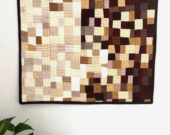 Puede incluir: Un edredón de pared rectangular que presenta una cuadrícula de pequeños parches de tela cuadrados en un degradado de colores que van desde el crema, beige y bronceado claro a la izquierda hasta tonos marrón oscuro, burdeos y chocolate.