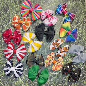 May include: A collection of colorful, decorative hair bows in various patterns. The bows feature stripes, polka dots, paisley, and seasonal designs, including pumpkins and fall leaves. The bows are made of ribbon and are suitable for pets or children.