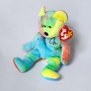 May include: A tie-dye blue, yellow, and orange beanie baby bear with a peace sign on its chest. The bear has a red Ty tag with a white star.