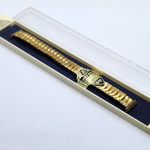 May include: A gold watch band with a gold and black shield-shaped emblem that says "Watch Band Swiss Precision 950". The band is in a white box with a clear plastic lid.