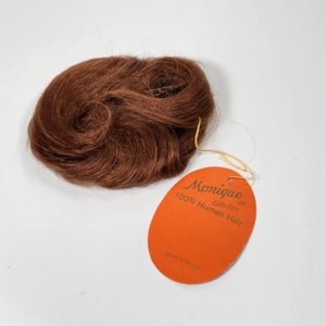 Puede incluir: Un pequeño paquete de cabello humano marrón con una etiqueta que dice "Monique Collection 100% Cabello Humano" y "Hecho en Malasia".