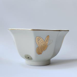 May include: White ceramic teacup with a gold rim and a gold butterfly design. The cup has a hexagonal shape.