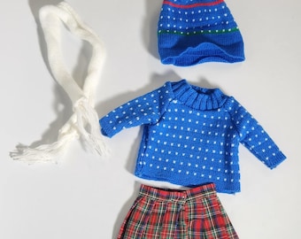 Könnte beinhalten: Ein winterliches Puppen-Outfit-Set. Das Set beinhaltet eine blaue Strickmütze mit weißem Bommel, einen blauen Pullover mit weißen Punkten, einen rot-grünen karierten Rock und einen weißen Schal.