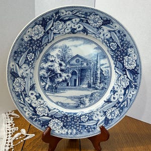 Puede incluir: Un plato redondo de cerámica con un patrón de transferencia azul y blanco. El centro representa un edificio clásico con columnas y árboles, mientras que el borde ancho está decorado con un denso motivo floral de rosas y hojas.