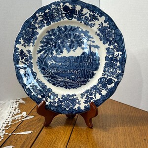 May include: A round ceramic plate featuring a blue and white transferware pattern. The center depicts a rural landscape with a church spire and trees, while the scalloped rim is decorated with intricate floral motifs and scrolling foliage.