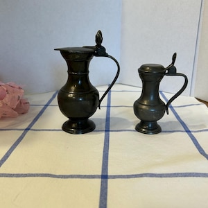 Puede incluir: Dos jarras de metal gris oscuro con tapas abatibles y asas curvas. La jarra más grande tiene un pico ancho, mientras que la más pequeña tiene un cuerpo más compacto y redondeado. Ambos recipientes se asientan sobre bases circulares.