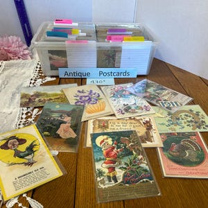 May include: A clear plastic storage bin filled with antique postcards, organized with colorful index tabs and labeled "Antique Postcards 470+". Several vintage postcards are displayed in front, featuring illustrations of Santa Claus, a turkey, and a hen.