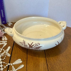 May include: A cream-colored ceramic serving bowl with two side handles and brown botanical illustrations on the exterior. A purple ruler is visible on the left, showing a measurement of approximately 9 inches across the bowl's diameter.