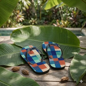 May include: A pair of flip-flops featuring a geometric pattern of squares in shades of navy blue, teal, and burnt orange. The footwear has black rubber thong straps and a flat sole design, showcasing a multi-colored grid aesthetic on the footbed.