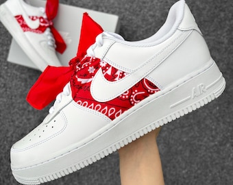 May include: White leather sneakers with a red bandana pattern on the side panels and a red ribbon tied around the laces. The sneakers have a white sole and the word "AIR" is visible on the side.