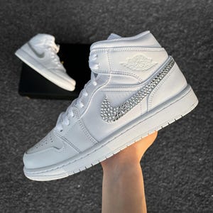 Könnte beinhalten: Weiße High-Top-Sneaker mit einem juwelenbesetzten Nike-Swoosh und juwelenbesetzten Akzenten an der Seite. Die Sneaker haben eine weiße Sohle und Schnürsenkel. Ein zweites Paar Sneaker ist im Hintergrund zu sehen.