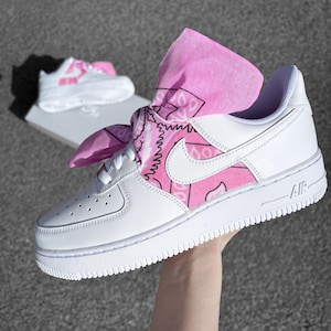 May include: White leather sneakers with a pink paisley design on the side and a matching pink bandana tied around the laces. The sneakers have a white sole and the word "AIR" printed on the side. Another pair of sneakers is visible in the background.