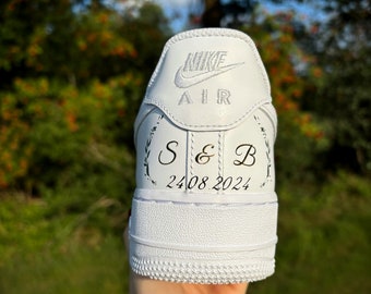 May include: White Nike Air Force 1 sneaker with custom lettering. The heel displays the Nike logo and the word "AIR". Below the logo are the initials "S & B" and the date "24.08.2024" in black script, framed by floral designs.