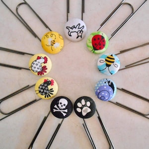 May include: Ten silver paper clips with colorful fabric buttons attached. The buttons feature various designs, including a bee, a ladybug, a chicken, a skull and crossbones, a paw print, a snail, and a cat.