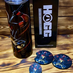 Puede incluir: Un vaso delgado de 20 onzas negro y rojo con un gr&aacute;fico de Deadpool sosteniendo una katana. El vaso est&aacute; empaquetado en una caja negra con el logotipo de HOGG y el texto "20 oz. Skinny Tumbler". La imagen tambi&eacute;n muestra tres posavasos redondos con el mismo gr&aacute;fico de Deadpool.