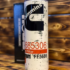 May include: White stainless steel tumbler with a black and white distressed graphic of the Cummins logo and the text "685306 Fuel Filter Guard FE5686".