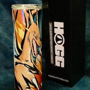 May include: A 20 oz. stainless steel skinny tumbler with a sublimation design featuring a cartoon image of a fiery orange and yellow Pokemon character. The tumbler is packaged in a black box with the text "HOGG" and "HOGGOUTFITTERS.COM".