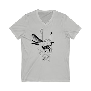 May include: A gray V-neck t-shirt with a black graphic of a hand making the rock and roll hand gesture. The hand is holding a pair of scissors and a comb. The hand is tattooed with a crescent moon, a rose, and other designs.
