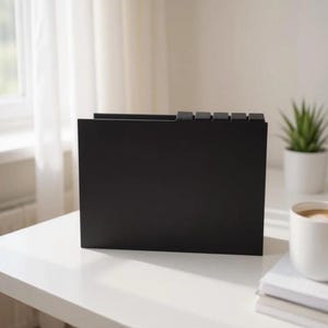 May include: A black file organizer with five tab dividers sits on a white table. A white coffee cup and a small potted plant are also on the table. The background includes a window and white curtains.