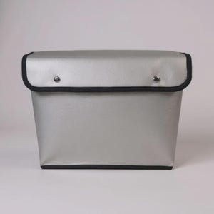 May include: A silver-colored storage box with a black trim around the edges. The lid is secured with two silver rivets. The box has a rectangular shape and a smooth, reflective surface. The overall design is simple and modern.