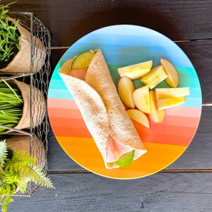 Peut inclure: Une assiette color&eacute;e avec un motif arc-en-ciel pr&eacute;sente un wrap au jambon et au concombre et des pommes en tranches.