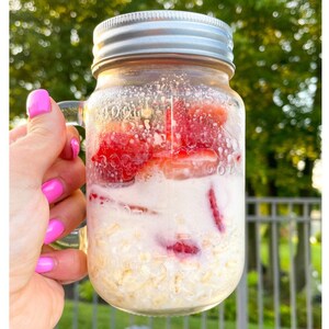 Peut inclure: Un bocal en verre rempli de flocons d'avoine, de fraises coup&eacute;es et de lait. Le bocal a un couvercle argent&eacute; et est tenu par une main avec du vernis &agrave; ongles rose.