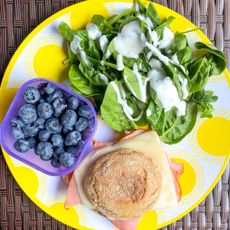 Peut inclure: Une assiette jaune avec un sandwich, une salade de feuilles de spinaches avec une vinaigrette cr&eacute;meuse et un r&eacute;cipient de myrtilles.