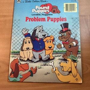 May include: A Little Golden Book titled "Pound Puppies: Problem Puppies". The cover features a group of cartoon puppies, including a grey puppy wearing a blue jacket, a yellow puppy with a red collar, and a brown puppy wearing a top hat and a red bow tie. The book is set against a blue background with a white fence.