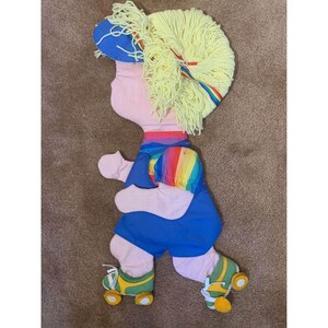May include: A vintage felt doll with yarn hair, wearing a rainbow striped shirt, blue pants, and yellow roller skates. The doll is in a skating pose.