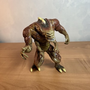 May include: A detailed action figure of a monstrous creature with a brown and green color scheme. The figure has a textured, reptilian appearance, with sharp teeth and claws. The figure is posed in a menacing stance.