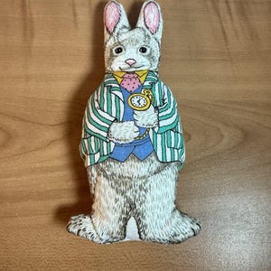 Może przedstawiać: Tkaninowa lalka królika ubrana w zielono-białą kurtkę w paski, niebieską kamizelkę i różowy krawat w kropki. Królik trzyma złoty zegarek kieszonkowy. Królik ma szare futro i różowe uszy.