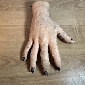 May include: A realistic-looking, pale-coloured, rubber hand with black fingernails. The hand is posed with fingers spread apart, as if reaching out.