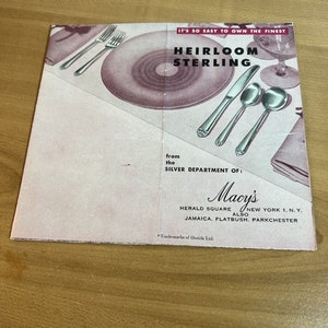 May include: A vintage advertisement for Heirloom Sterling silverware from Macy's. The brochure features a pink and white color scheme with images of silverware, plates, and glassware. Text includes "It's so easy to own the finest" and Macy's store locations.