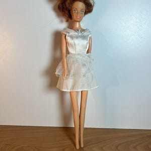 Puede incluir: Una muñeca vintage con cabello castaño y ojos azules que lleva un vestido blanco con una superposición de encaje.